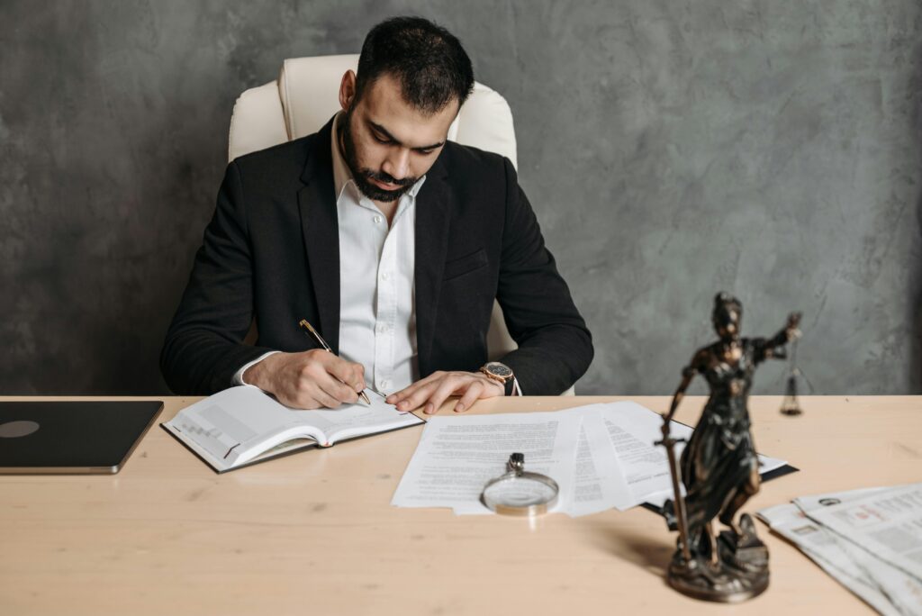 Attorney in formal suit writing notes at a desk with legal documents, laptop, and Lady Justice statue.